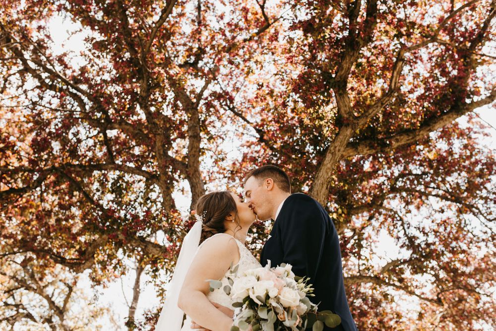 Bride and groom share a romantic kiss beneath a stunning red-leafed tree, bride holding elegant white floral bouquet.
