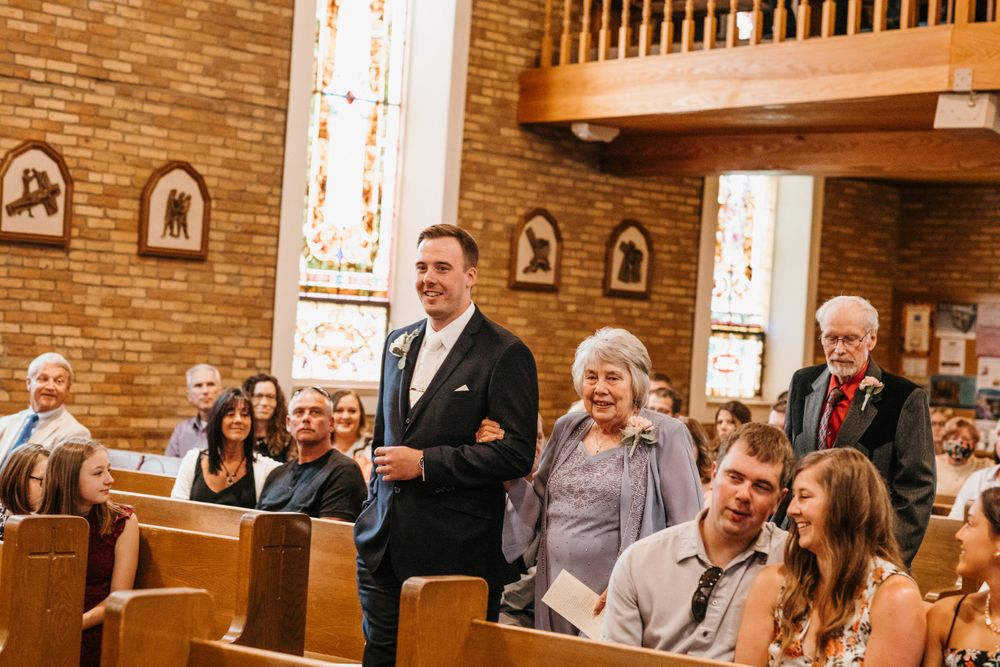 Groom escorts elderly woman down church aisle past seated wedding guests in warm brick interior church.