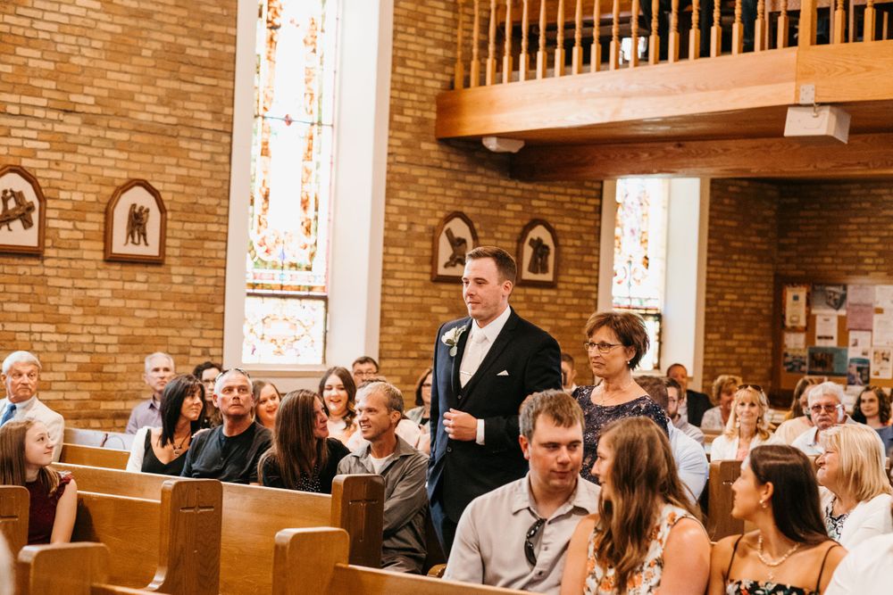 Groom walks elderly guest down the aisle of a traditional brick church filled with seated wedding attendees.