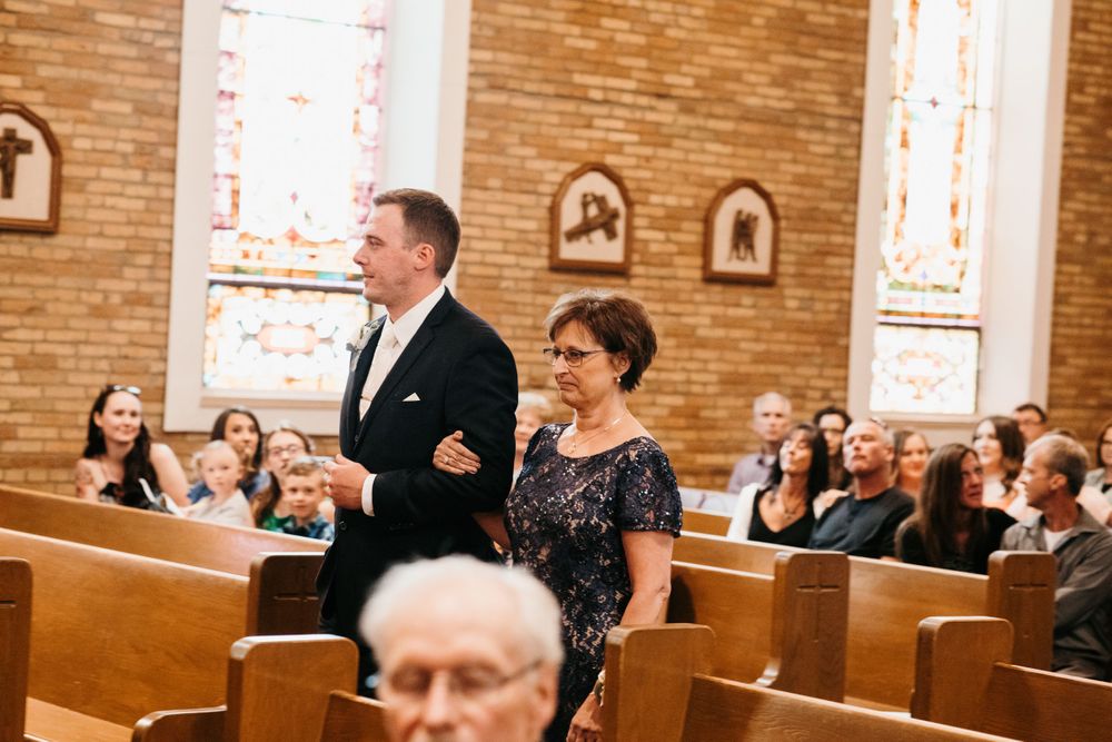 Groom escorts a woman in a floral dress down the aisle of a brick church with stained glass windows.