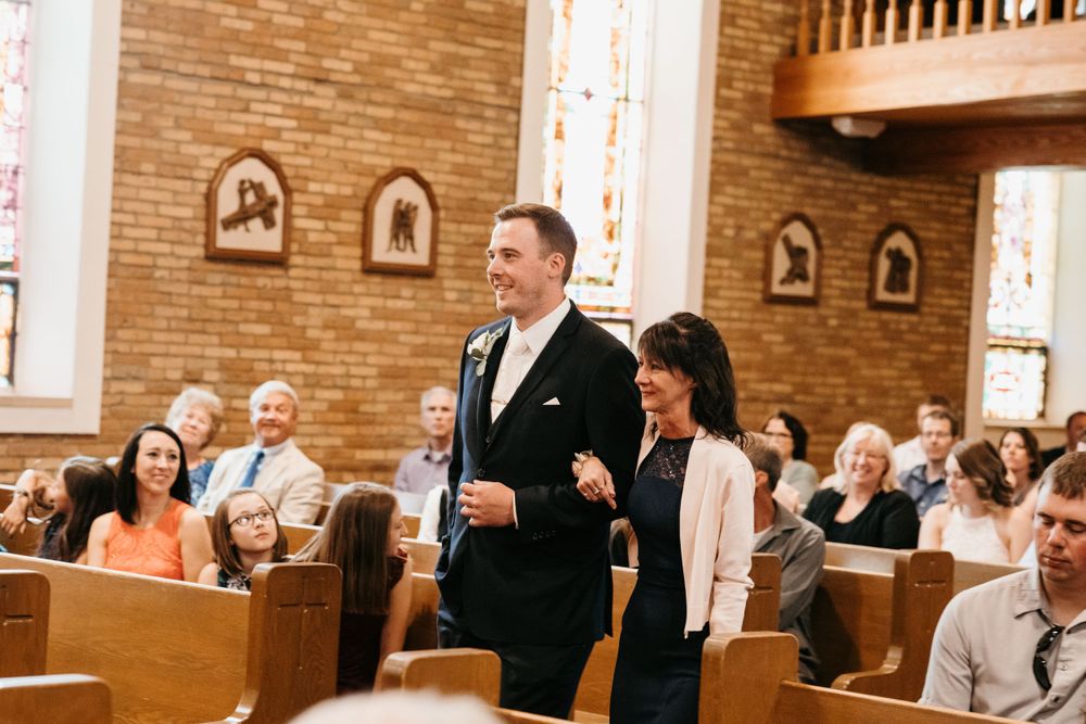 A smiling groom in a black tuxedo walks down the church aisle with a woman during a wedding ceremony.
