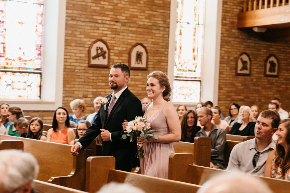 A bridesmaid in a pink dress carries flowers while walking down the church aisle with a groomsman.