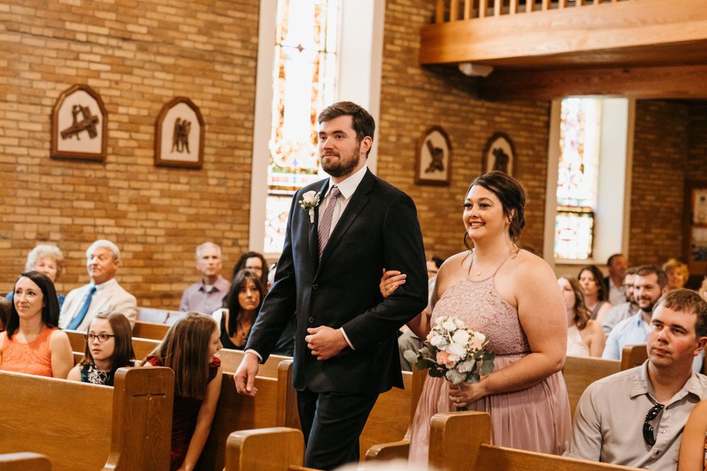 A groomsman escorts a smiling bridesmaid holding a bouquet down the aisle of a brick church.