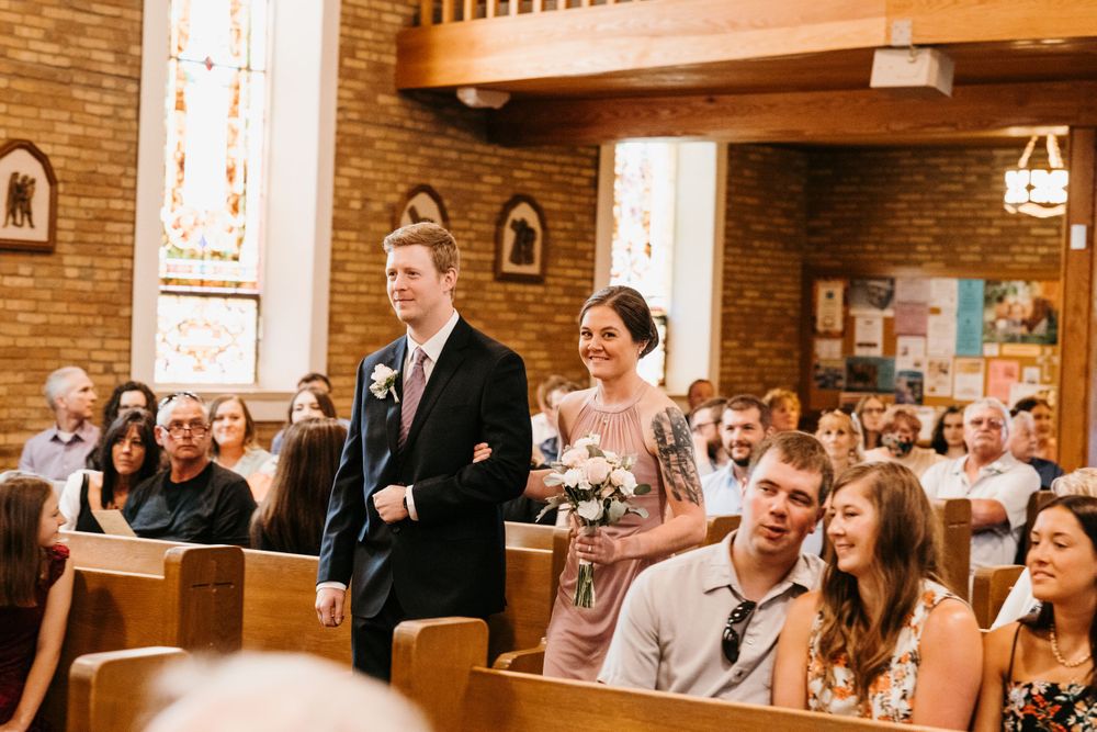 A groomsman in a dark suit walks a smiling bridesmaid with flowers down a church aisle.
