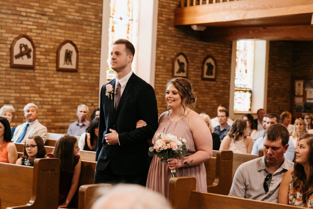 A bridesmaid in a blush gown carries a floral bouquet while being escorted down the church aisle.