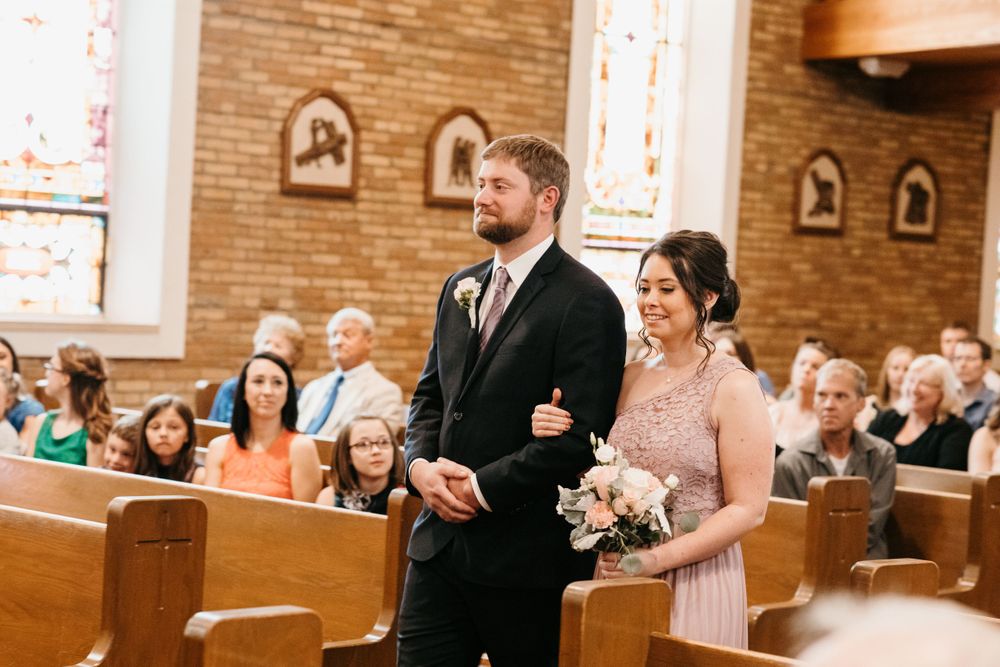 A smiling bridesmaid in a pink dress holds a bouquet while walking down the aisle with a groomsman.