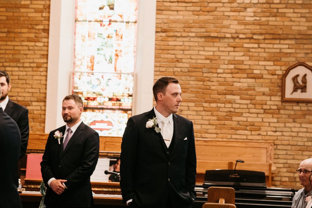 A groom in a black suit stands at the altar with groomsmen beside a stained glass church window.