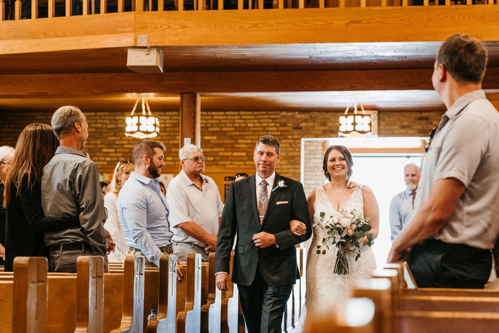 A beaming bride in white holds a bouquet while being walked down the wooden church aisle by her father.