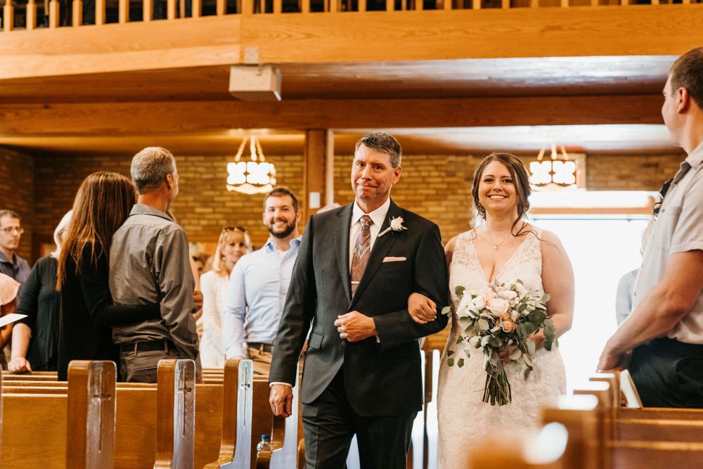 A happy bride carrying a greenery bouquet walks down the church aisle with her escort during the ceremony.