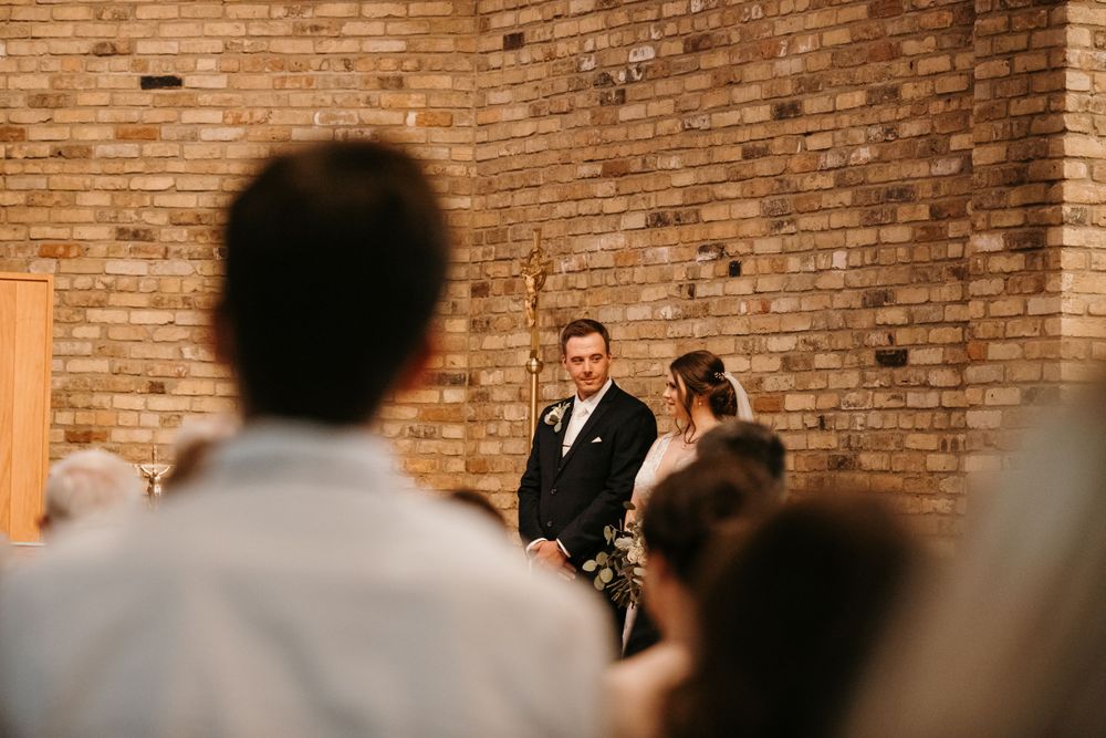 Groom stands at the altar watching guests during a wedding ceremony in a brick-walled church.
