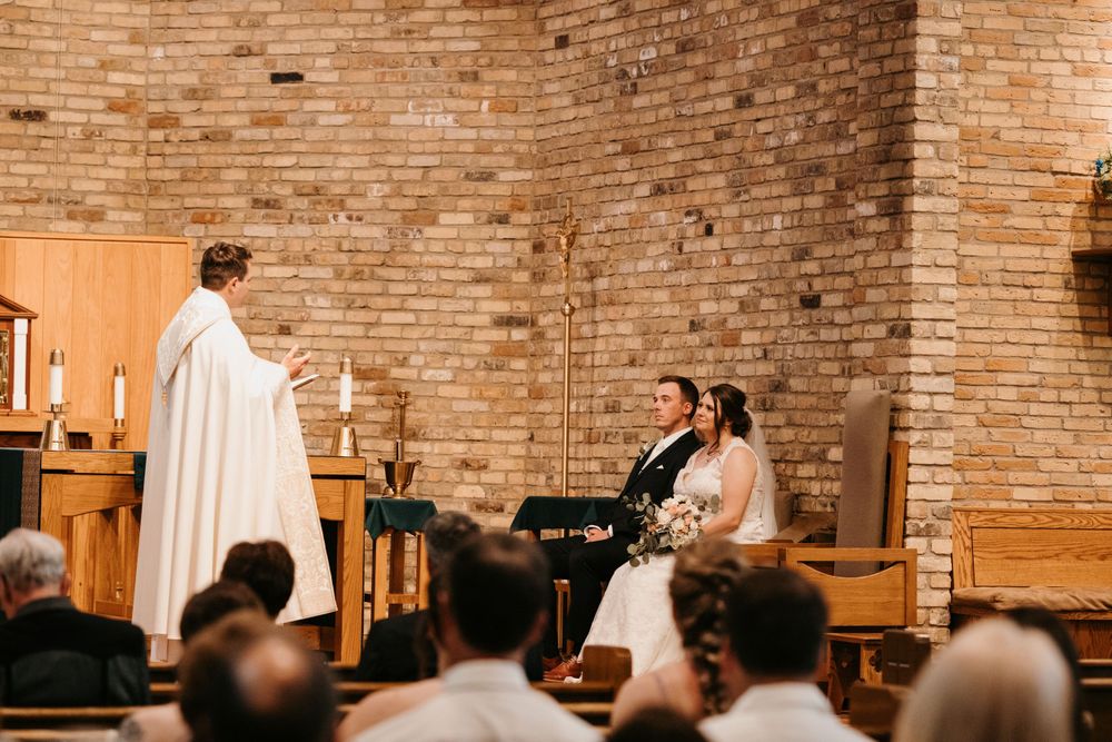 Officiant in white robes addresses bride and groom seated at the altar during a church wedding ceremony.