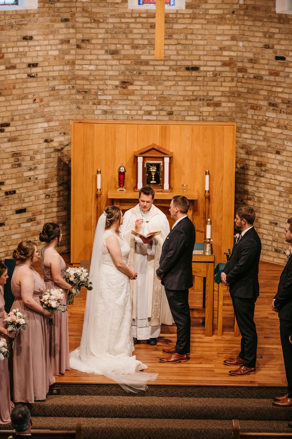 Bride and groom exchange vows with officiant at a wooden altar surrounded by bridesmaids and groomsmen.