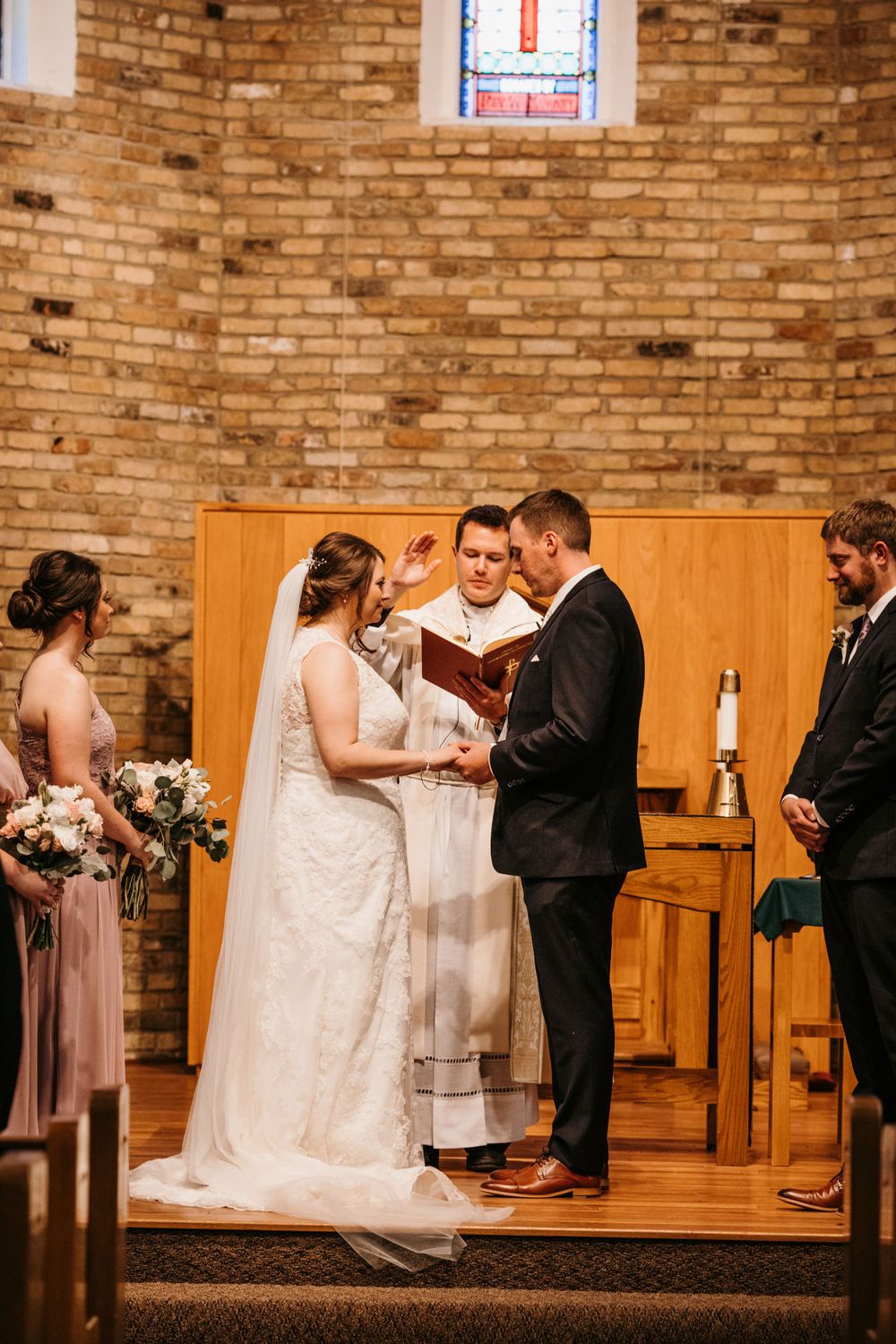 Officiant blesses bride and groom during vow exchange at a wooden altar in a brick church.