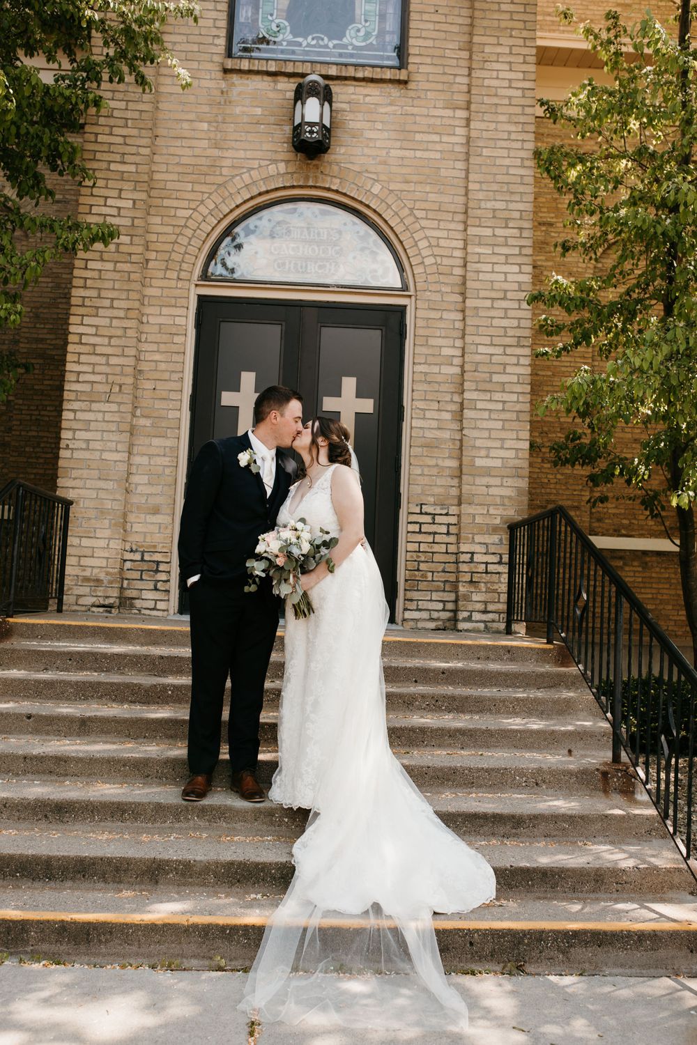 Bride and groom kiss on the steps outside a brick church with veil flowing in the breeze.