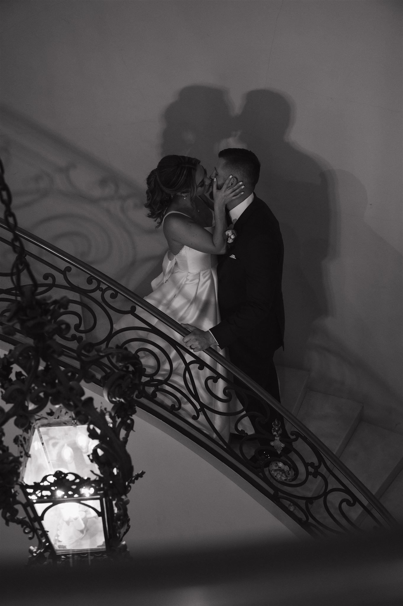 A romantic silhouette of newlyweds sharing a kiss on an elegant staircase with ornate wrought iron railings.