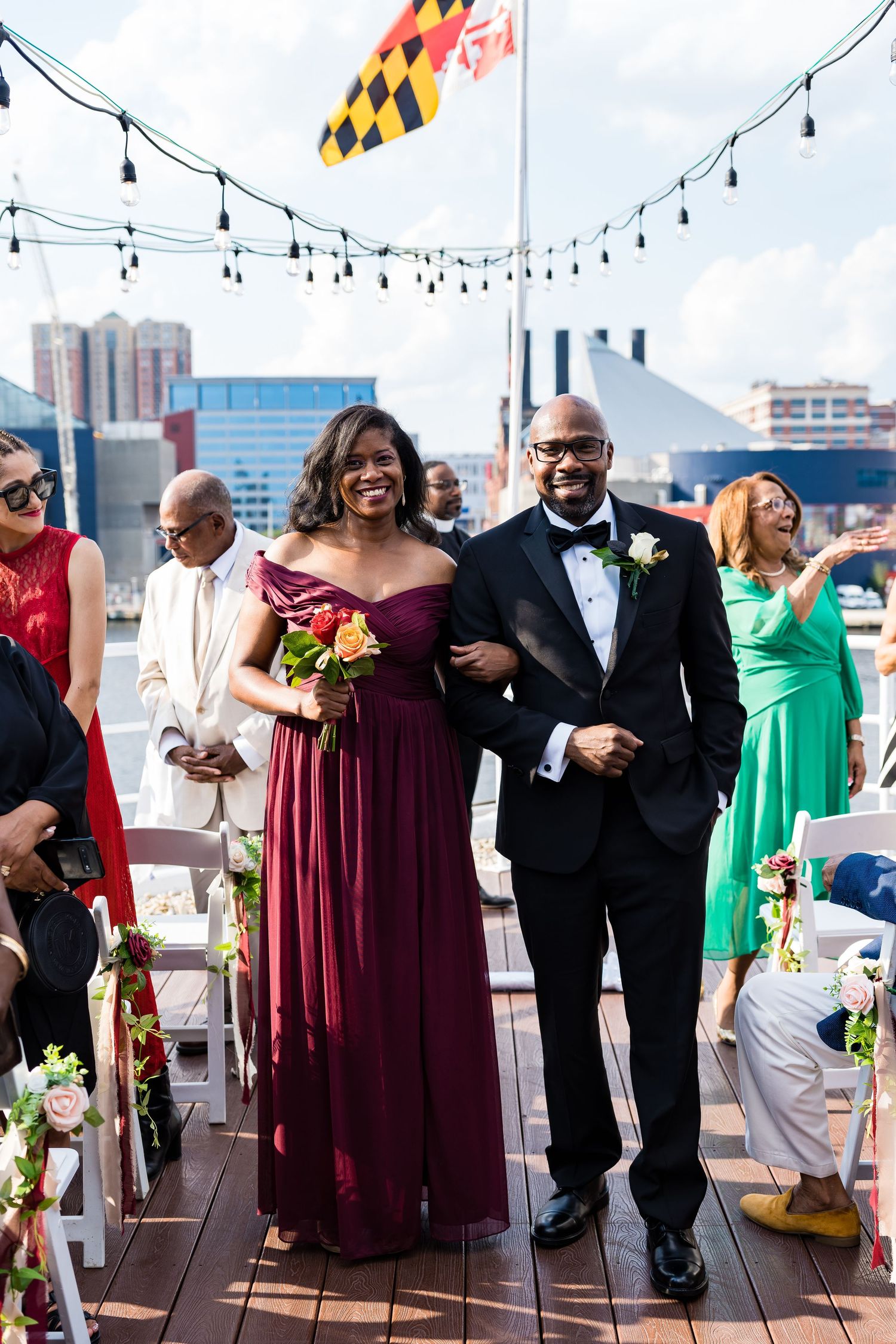 Wedding party members walking down the aisle under string lights with Maryland flag flying overhead.