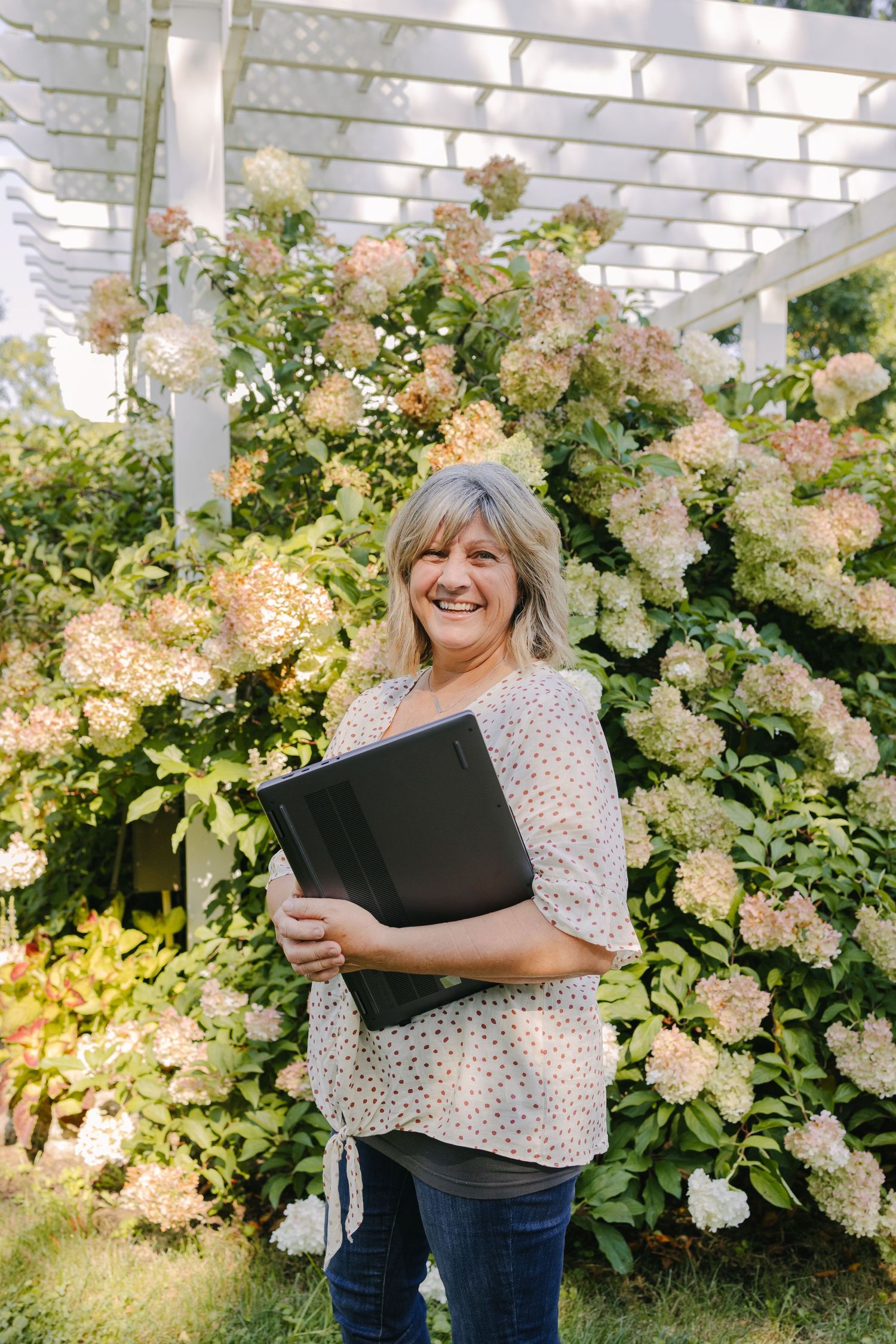 Person holding a portfolio while standing in front of blooming hydrangea bushes under a pergola.