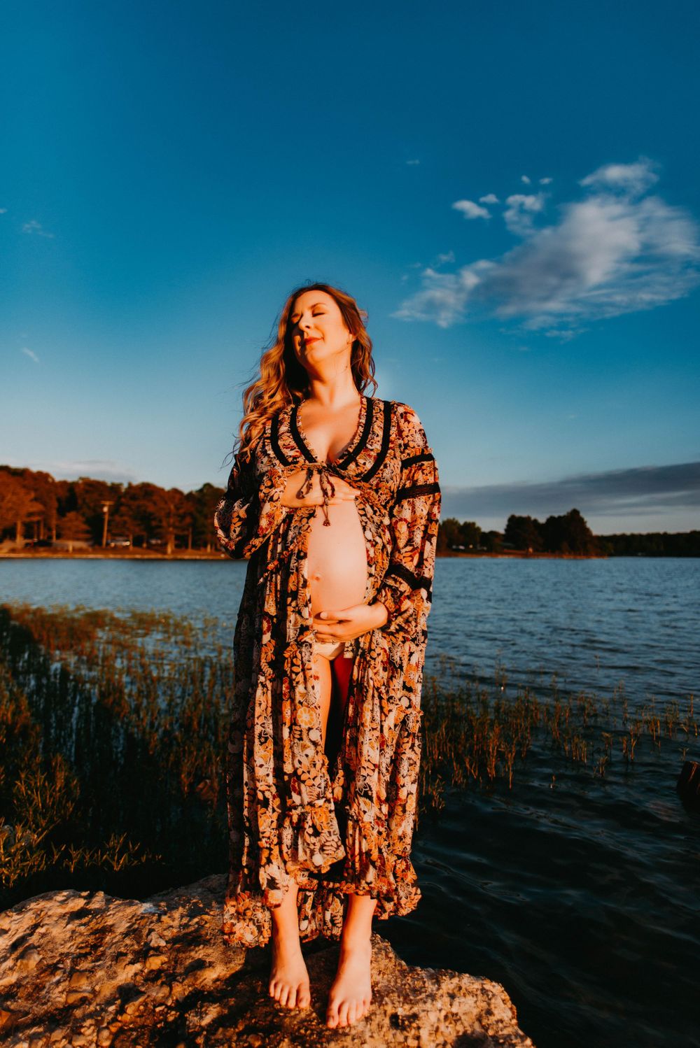 Silhouette of person in patterned dress against lakeside sunset with warm golden light.
