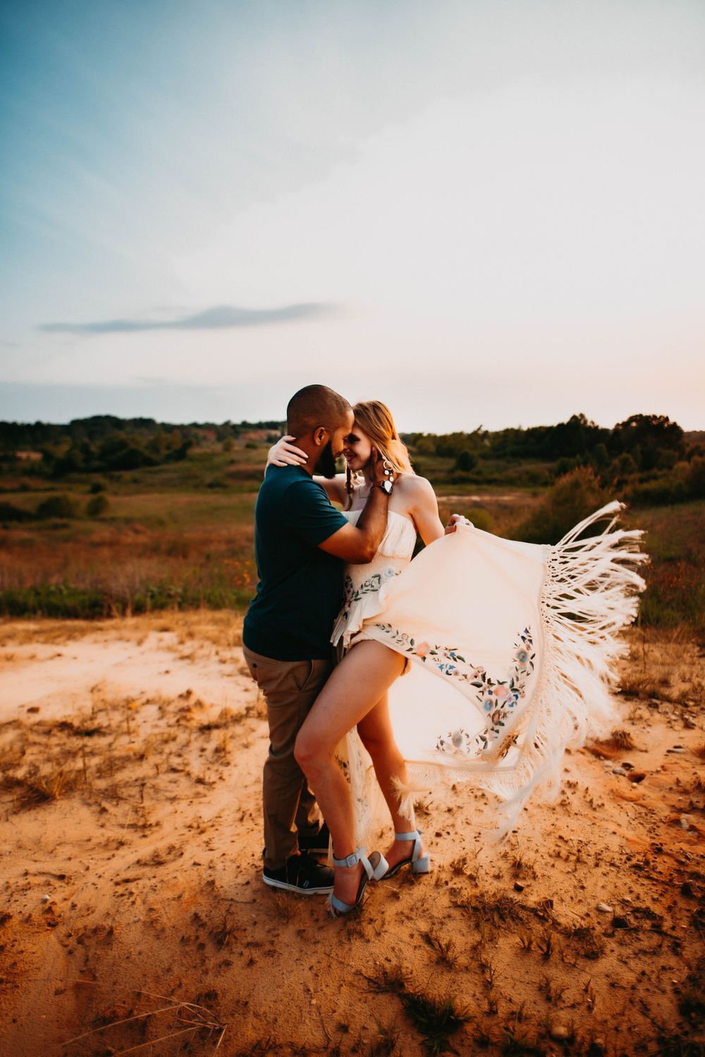 Couple embraces on sandy hill as white dress flows in wind during sunset.