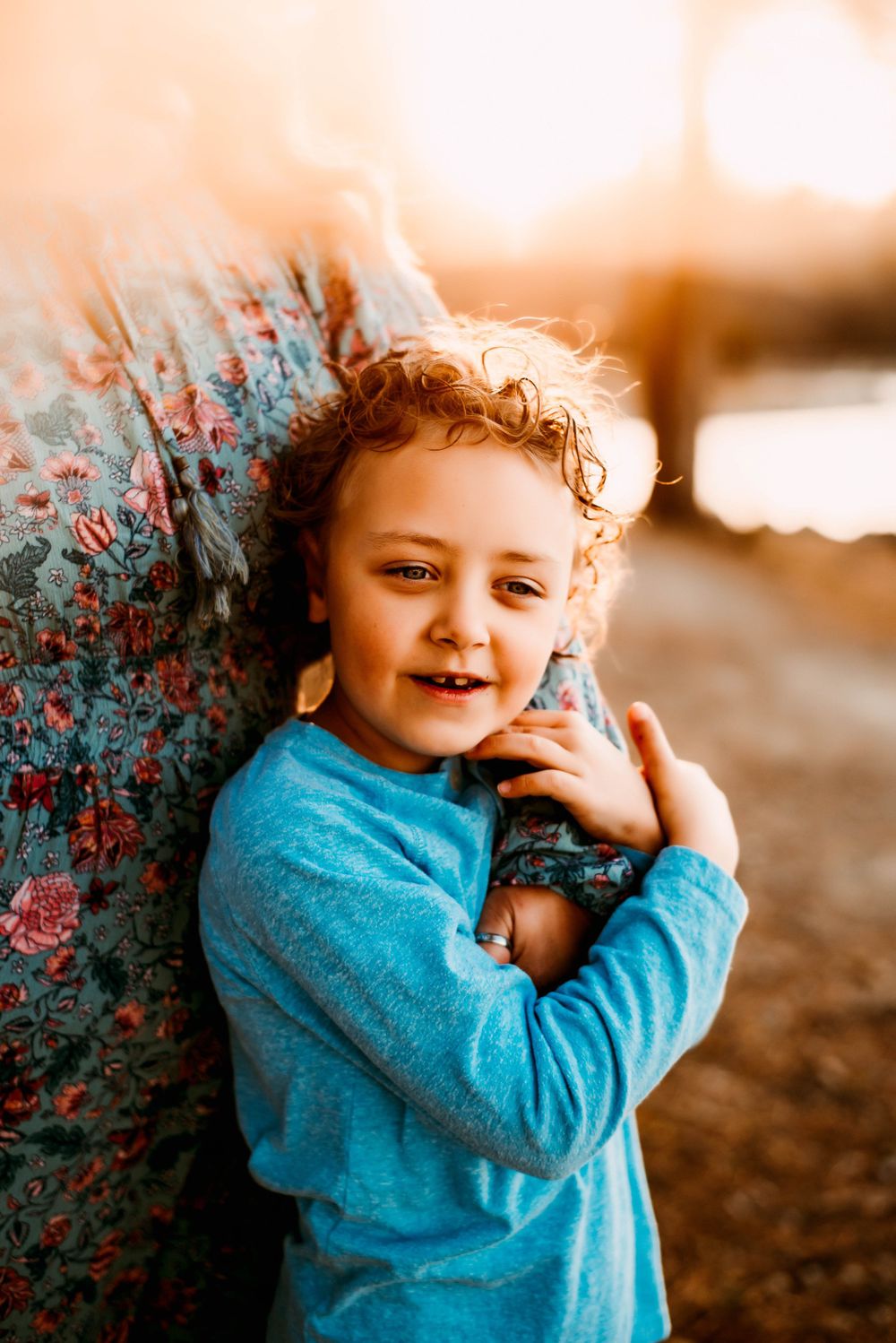 A young child in a blue sweater leans against a textured wall with red berries at sunset.