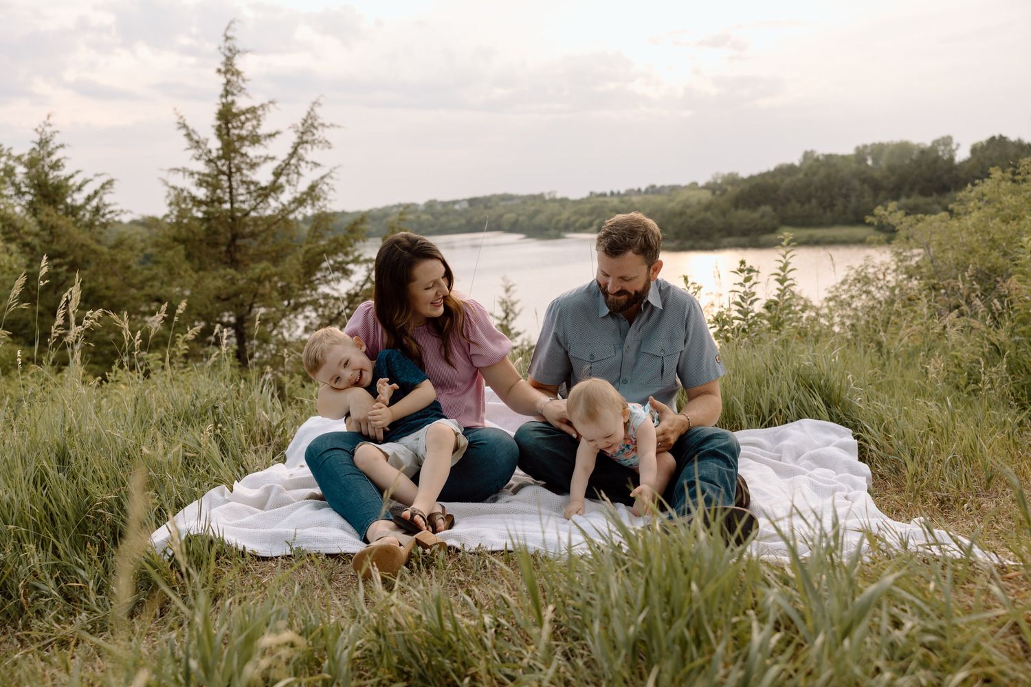Couple relaxing on picnic blanket with dog beside lake during sunset.