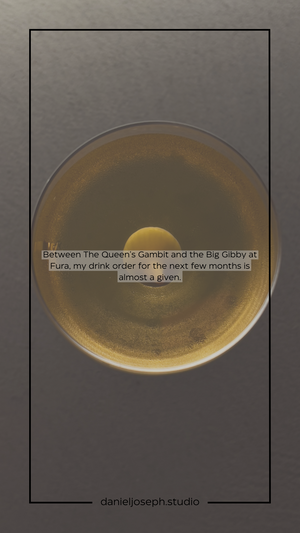A golden cocktail with a sphere garnish in a coupe glass against a gray background.
