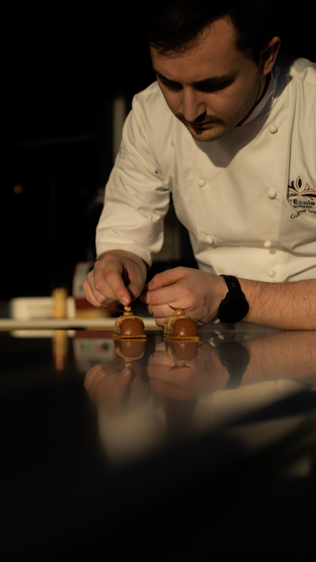 A chef in white uniform carefully decorates chocolate desserts on a dark countertop.