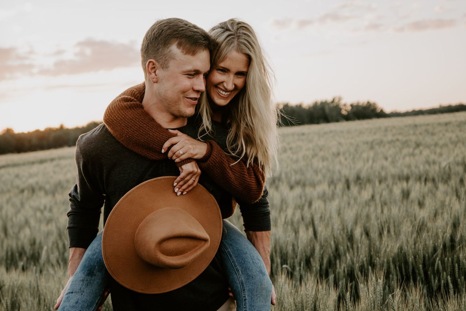 woman piggy backing on man holding fedora hat in hayfield