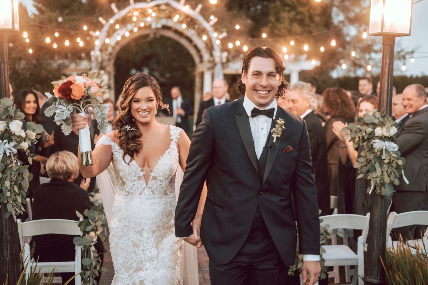 Newlyweds walk hand in hand down the aisle after their outdoor wedding ceremony under string lights.