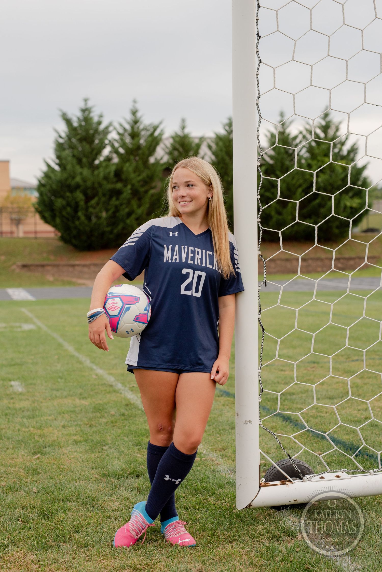 Soccer player in navy uniform poses with ball by goal post on green field.