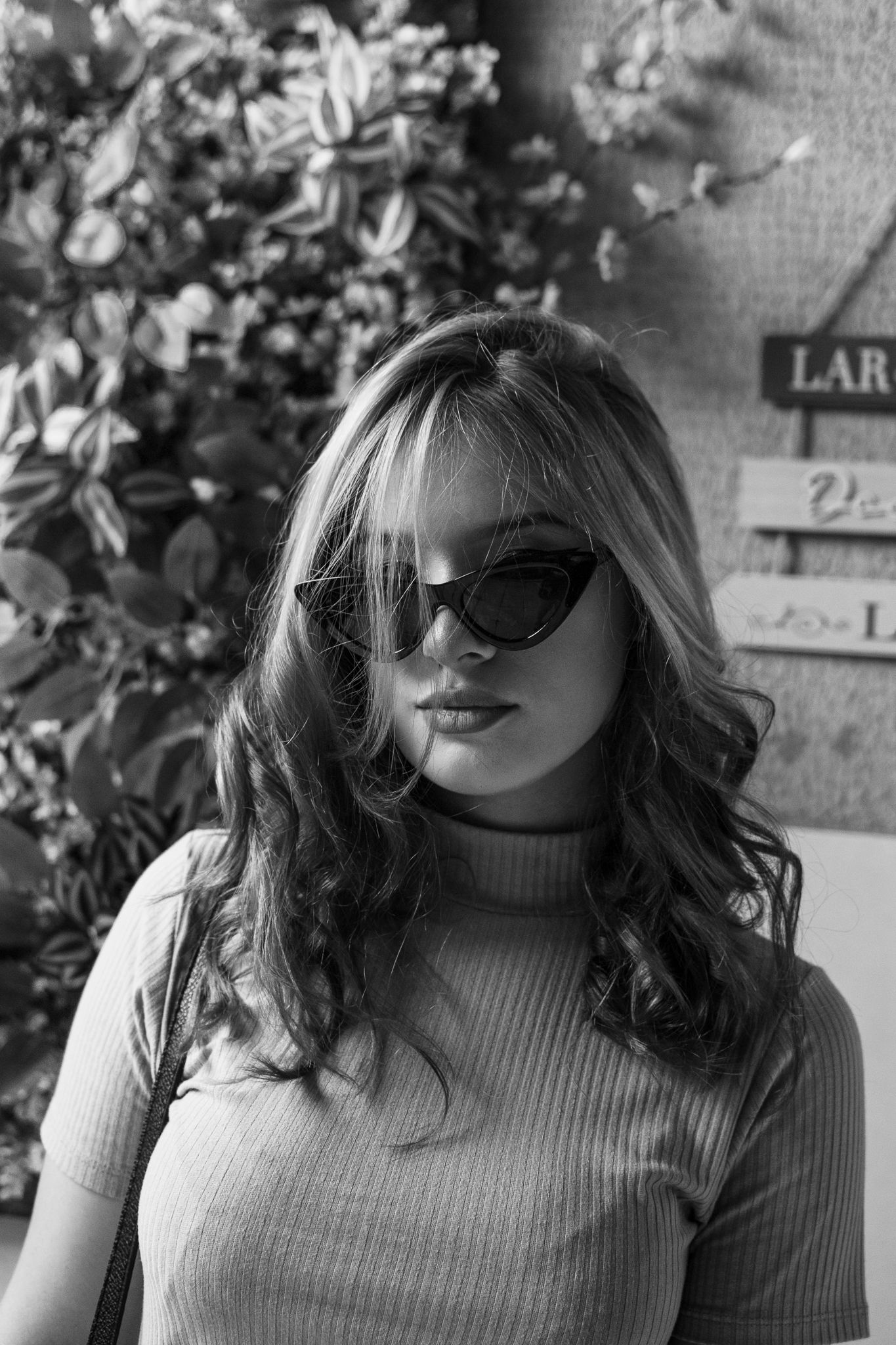 Black and white portrait with sunglasses and turtleneck sweater against foliage background.