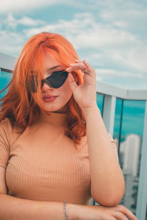 Person with vibrant orange hair wearing sunglasses and beige top poses against a turquoise cityscape backdrop.