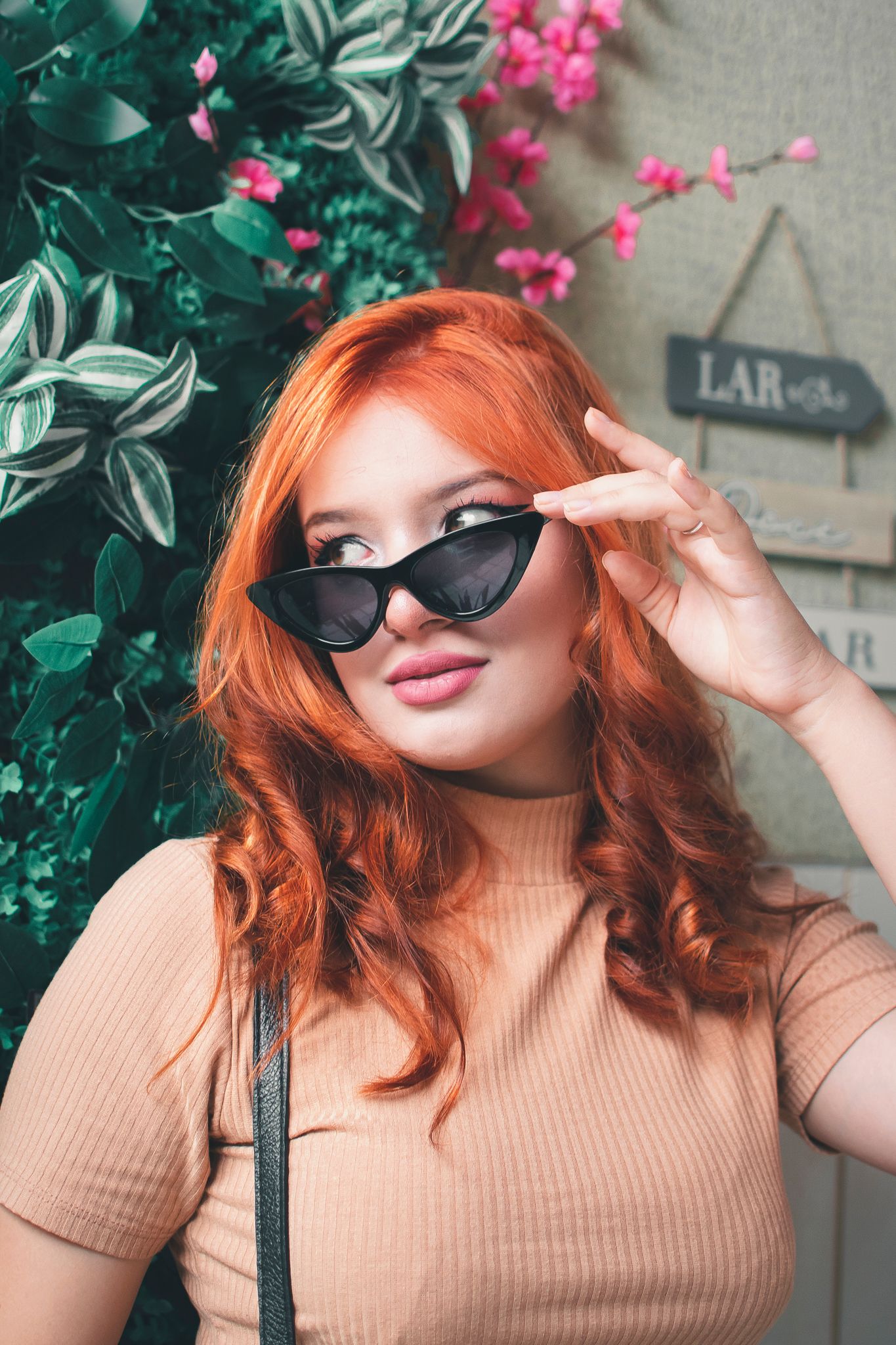 Stylish fashion portrait with cat-eye sunglasses against pink flowers and green foliage backdrop.