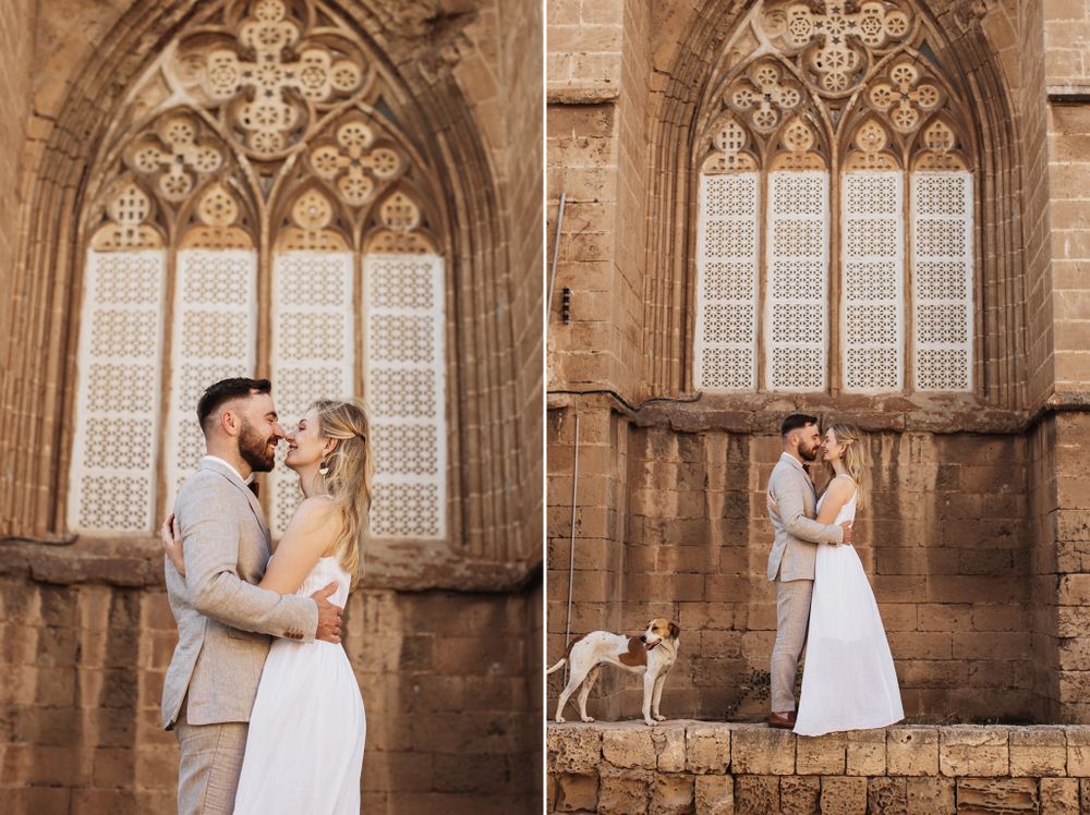 Romantic wedding couple embracing near gothic stone church windows with a white dog beside them.