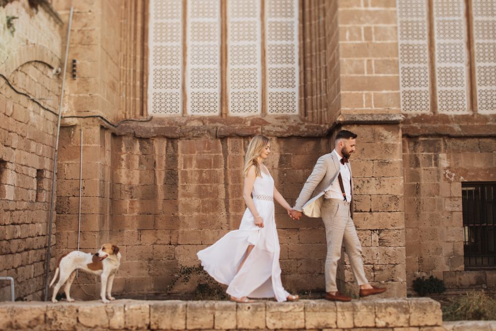 Bride and groom walk hand in hand past a rustic stone building with a white dog following them.