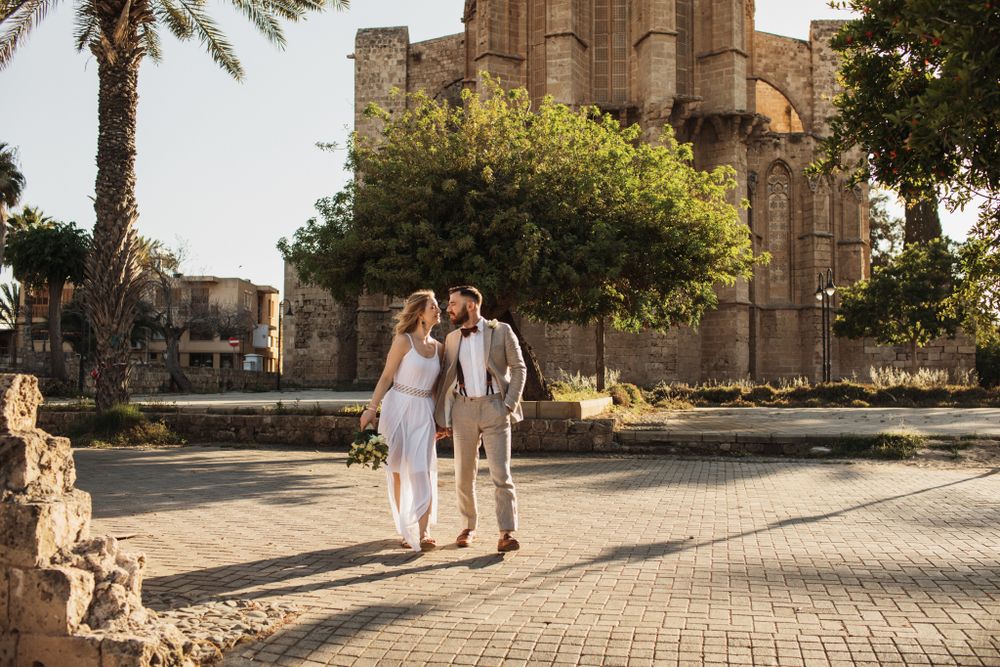 Wedding couple strolls through a sunlit plaza with an ancient stone cathedral in the background.
