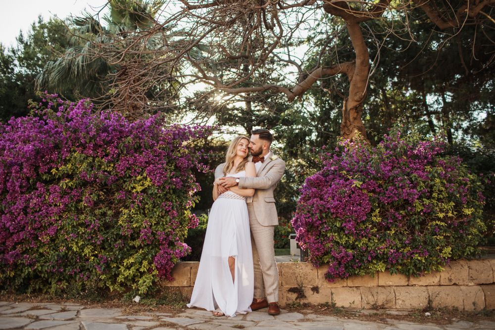 Bride and groom share a romantic embrace in front of lush purple flowering bougainvillea bushes.
