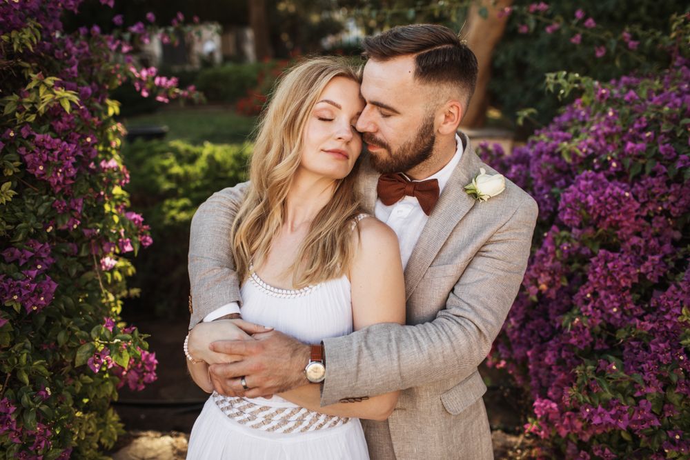 Groom tenderly embraces bride surrounded by vibrant purple bougainvillea flowers in a garden.