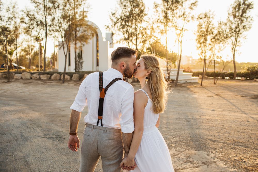 Newlyweds share a golden sunset kiss near a small white chapel in an arid landscape.
