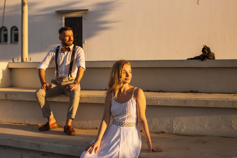 Groom sits on white steps above bride posing separately in warm evening golden hour light.