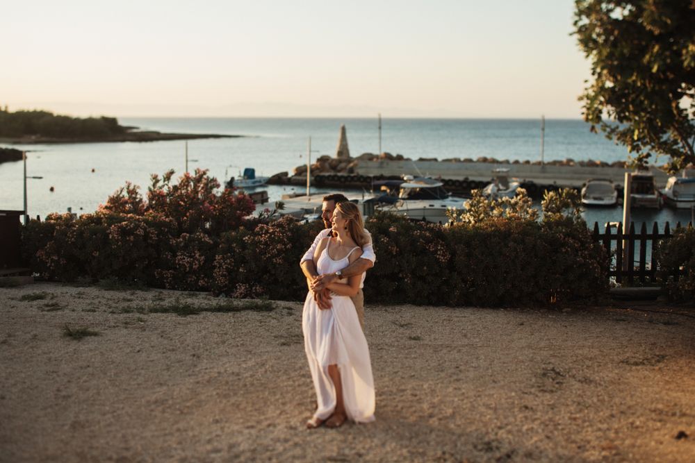 Bride in flowing white dress stands near a scenic harbor with the sea glowing at sunset.