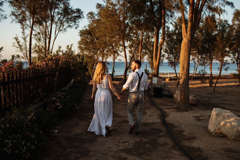 A couple in wedding attire walks hand in hand through a tree-lined path near the ocean at golden hour.