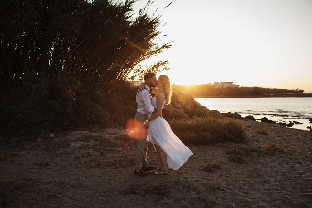 A bride and groom share a romantic kiss on a sandy beach at sunset with warm golden light.
