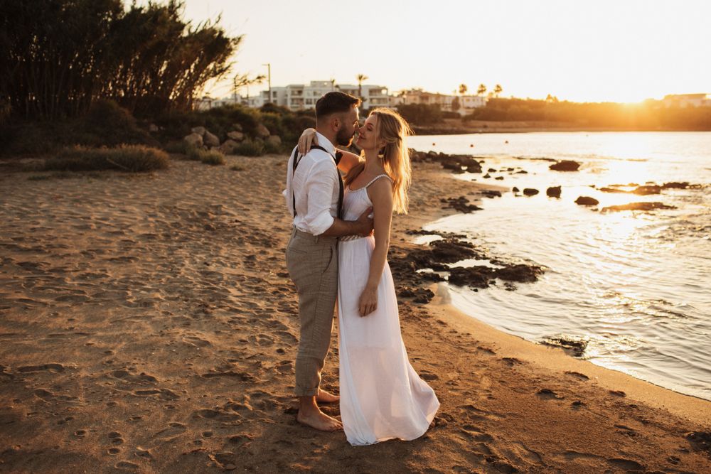 A newlywed couple embraces on a sandy beach at sunset with rocky shoreline and buildings in background.