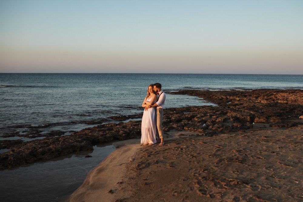 A wedding couple embraces on a rocky beach at dusk with calm blue ocean stretching to the horizon.