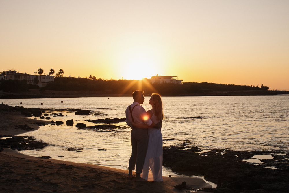 A bride and groom stand silhouetted together on a beach during a stunning golden sunset over the sea.