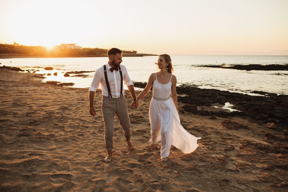 A smiling bride and groom walk hand in hand along a sandy beach during a beautiful golden sunset.