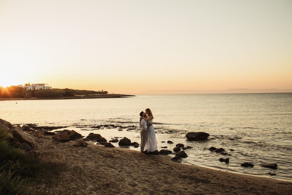 A wedding couple shares a tender embrace on a sandy beach during a soft pastel sunset by the sea.