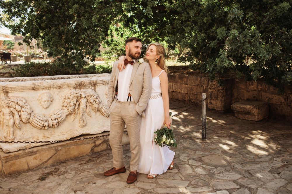 Bride and groom posing near ornate stone fountain in a sunlit Mediterranean garden.