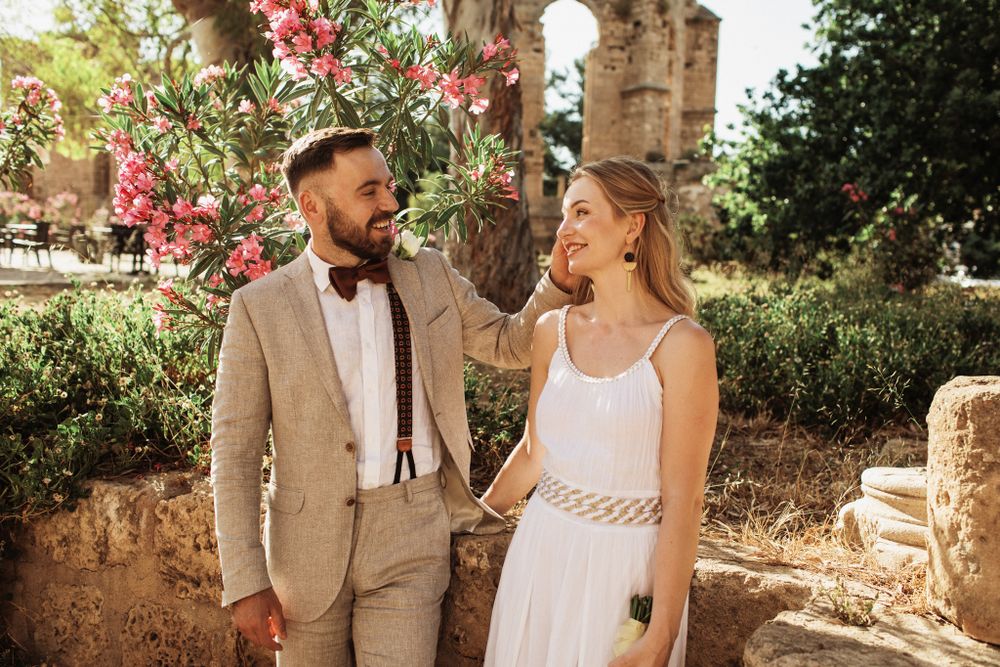 Smiling wedding couple holding hands near pink flowering oleander at ancient ruins.